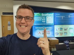 A smiling man wearing glasses and a navy blue jumper takes a selfie at DATA: Scotland 2025, pointing towards the conference information screens behind him.
