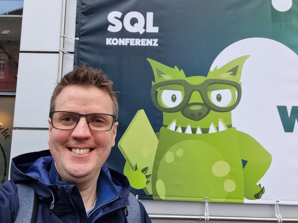 A smiling man wearing glasses and a navy blue jacket takes a selfie in front of a SQL Konferenz banner featuring a large green cartoon monster wearing glasses.