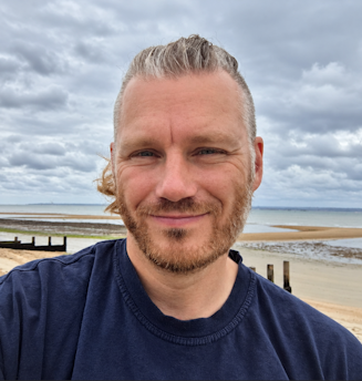 A smiling man with tied-back blond hair and a short beard takes a selfie on a sandy beach, wearing a navy blue T-shirt. Behind him are wooden groynes, the sea and a cloudy sky.