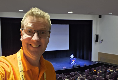 A smiling man wearing glasses, an orange polo shirt and a conference lanyard takes a selfie from the balcony of an auditorium, overlooking rows of seats and a speaker presenting on stage.